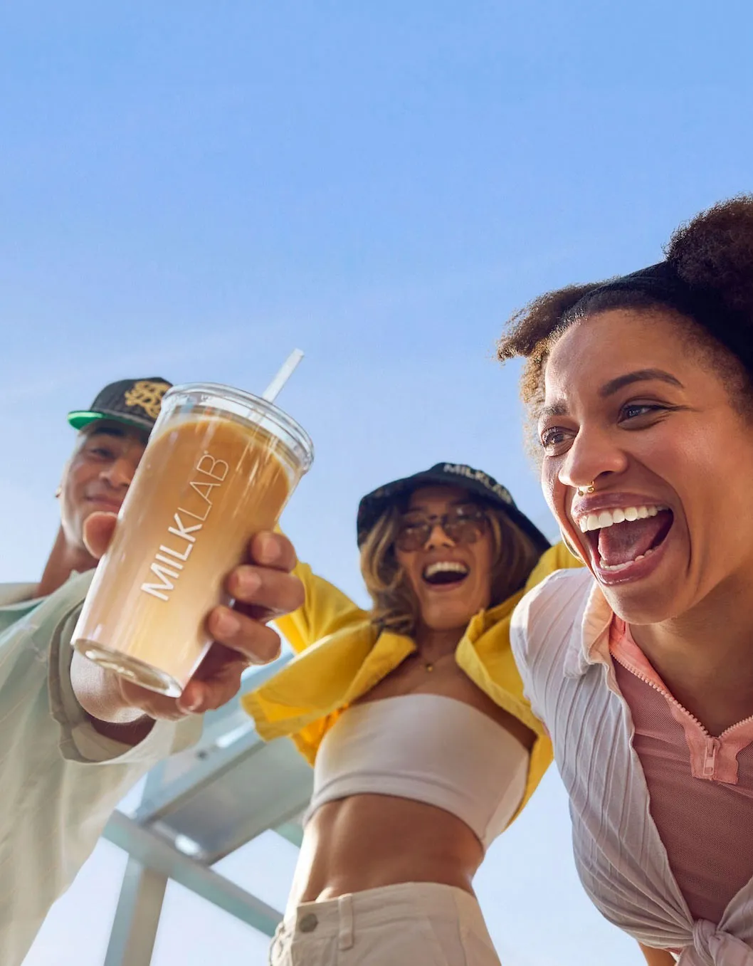 Three smiling young people outdoors, one holding a cup labeled MILKLAB with iced coffee against a clear blue sky.