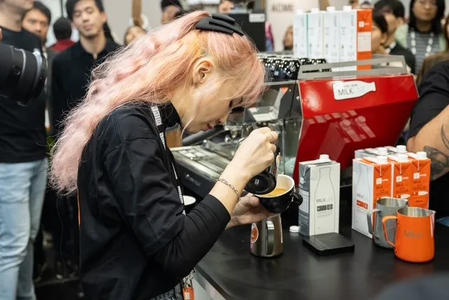 Barista with pink hair pouring milk into a cup at a coffee machine during a demonstration with onlookers nearby.