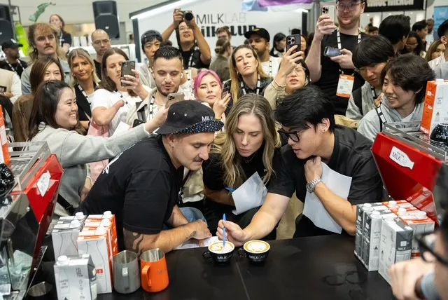 A group of people closely watching a latte art demonstration at a coffee event, with two cups of latte on the table and the demonstrator using a tool on the foam.