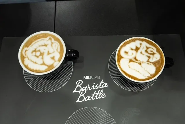 Two cups of latte art coffee with intricate designs placed on a table with 'MILKLAB Barista Battle' text between them.