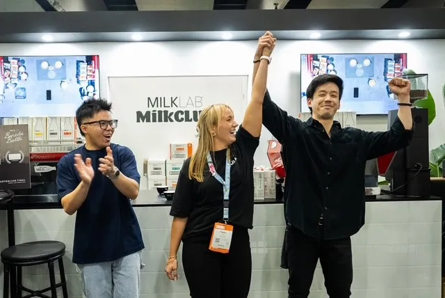 Three people standing indoors with two holding hands raised in celebration while the third applauds.