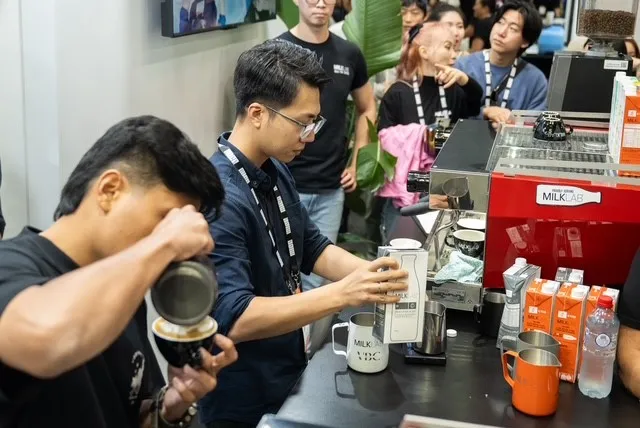 Two men preparing coffee drinks behind a counter with a red MILKLAB espresso machine while several people watch in the background.