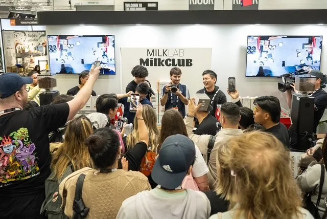 Crowd of people taking photos and videos of three men smiling and posing in front of a MILKLAB MilkClub display with two screens showing overhead footage.
