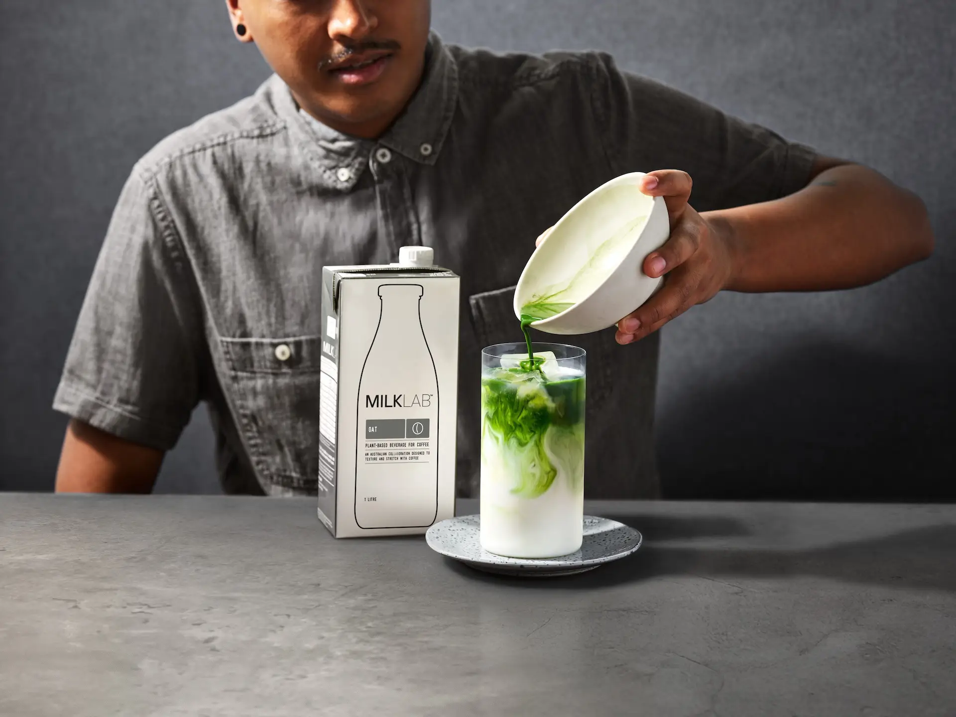 Person pouring green matcha tea into a glass of oat milk with Milklab oat milk carton on the table.