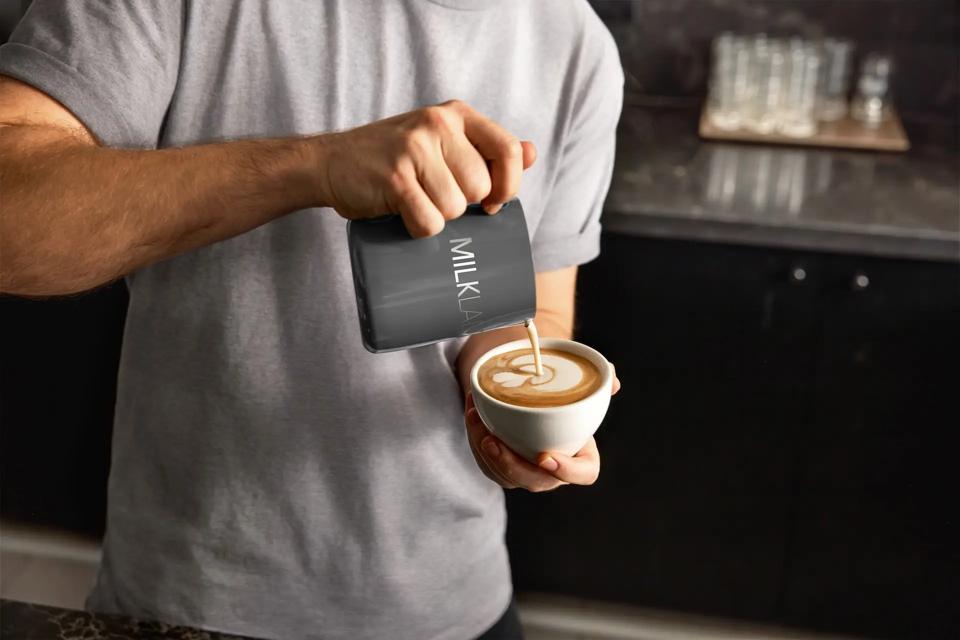 Person pouring milk from a small black jug labeled 'MILK' into a cup of coffee creating latte art.