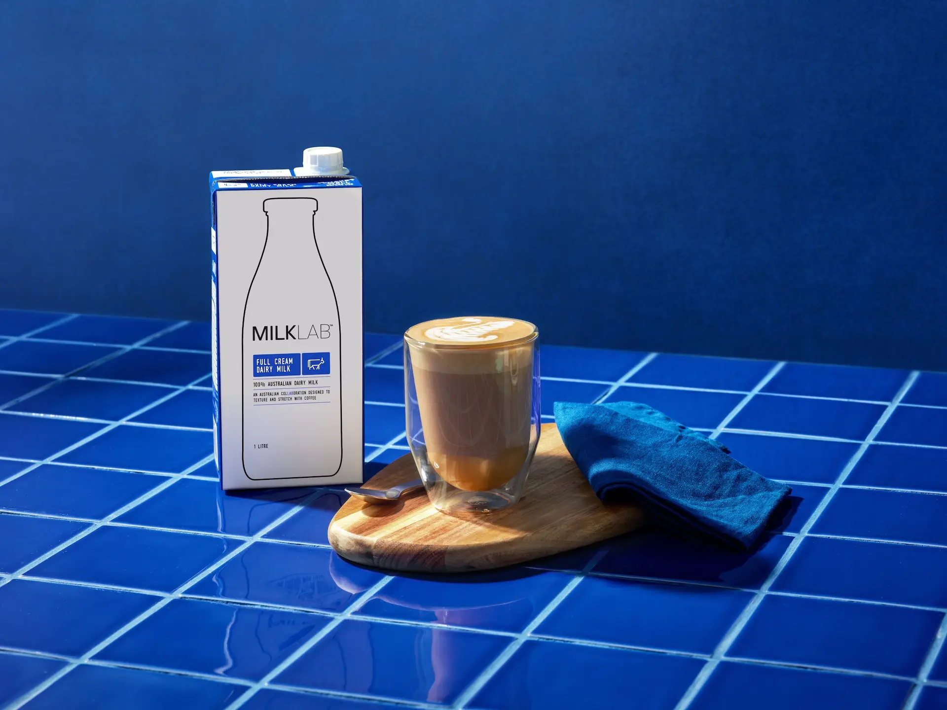 Carton of MilkLab full cream dairy milk next to a double-walled glass of latte with latte art, on a wooden board with a blue napkin and spoon, on a blue tiled surface.