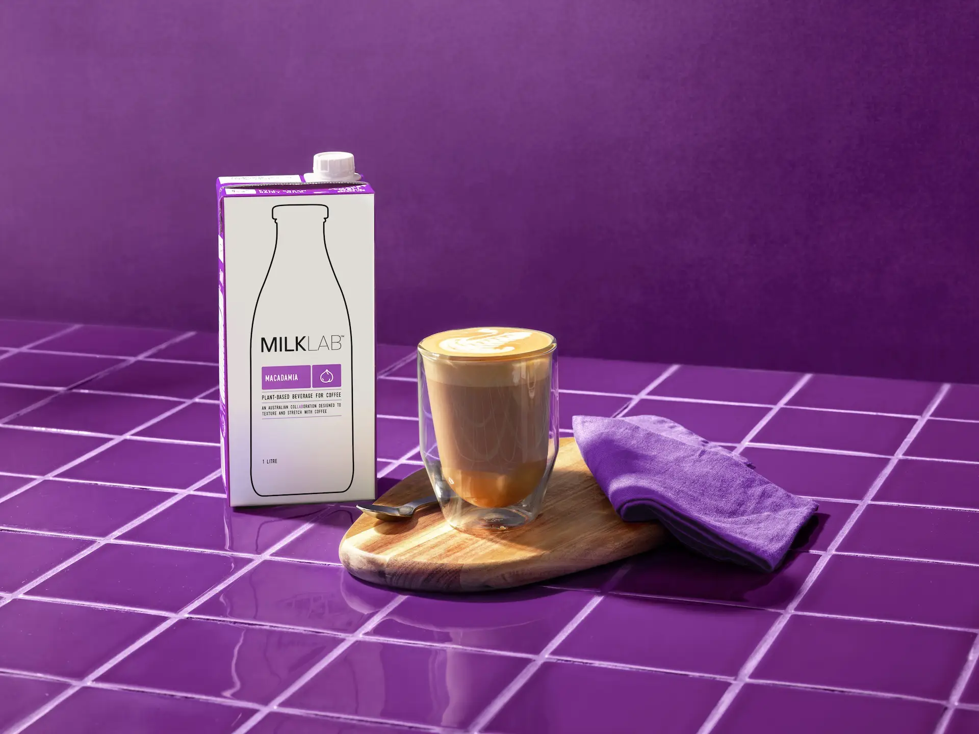 Carton of MilkLab macadamia plant-based beverage next to a latte with foam art on a wooden board, with a purple napkin on a purple tiled surface.