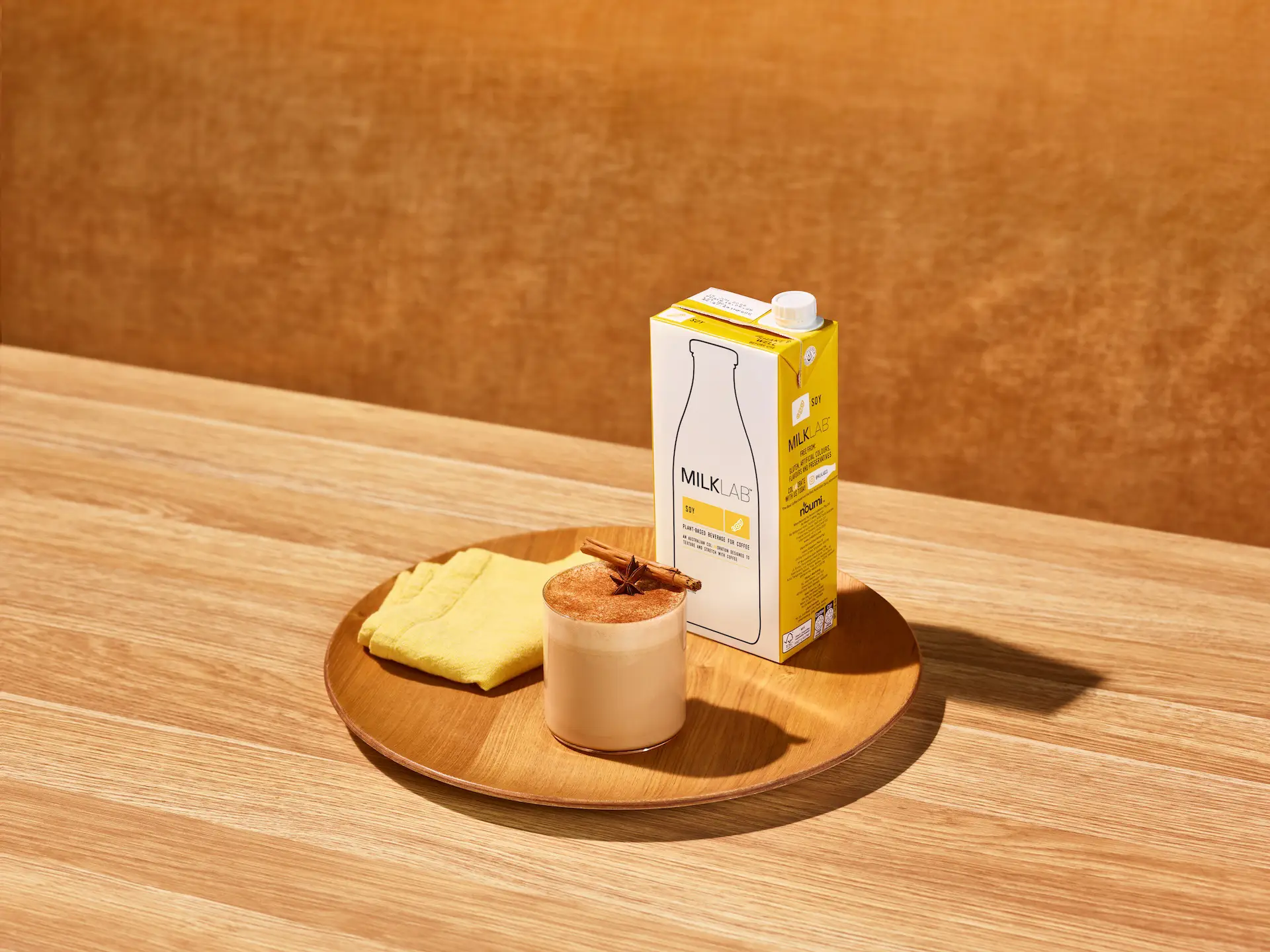 Wooden tray on a table holding a carton of MILKLAB soy plant-based milk, a glass of frothy drink topped with cinnamon stick and star anise, and a folded yellow cloth.