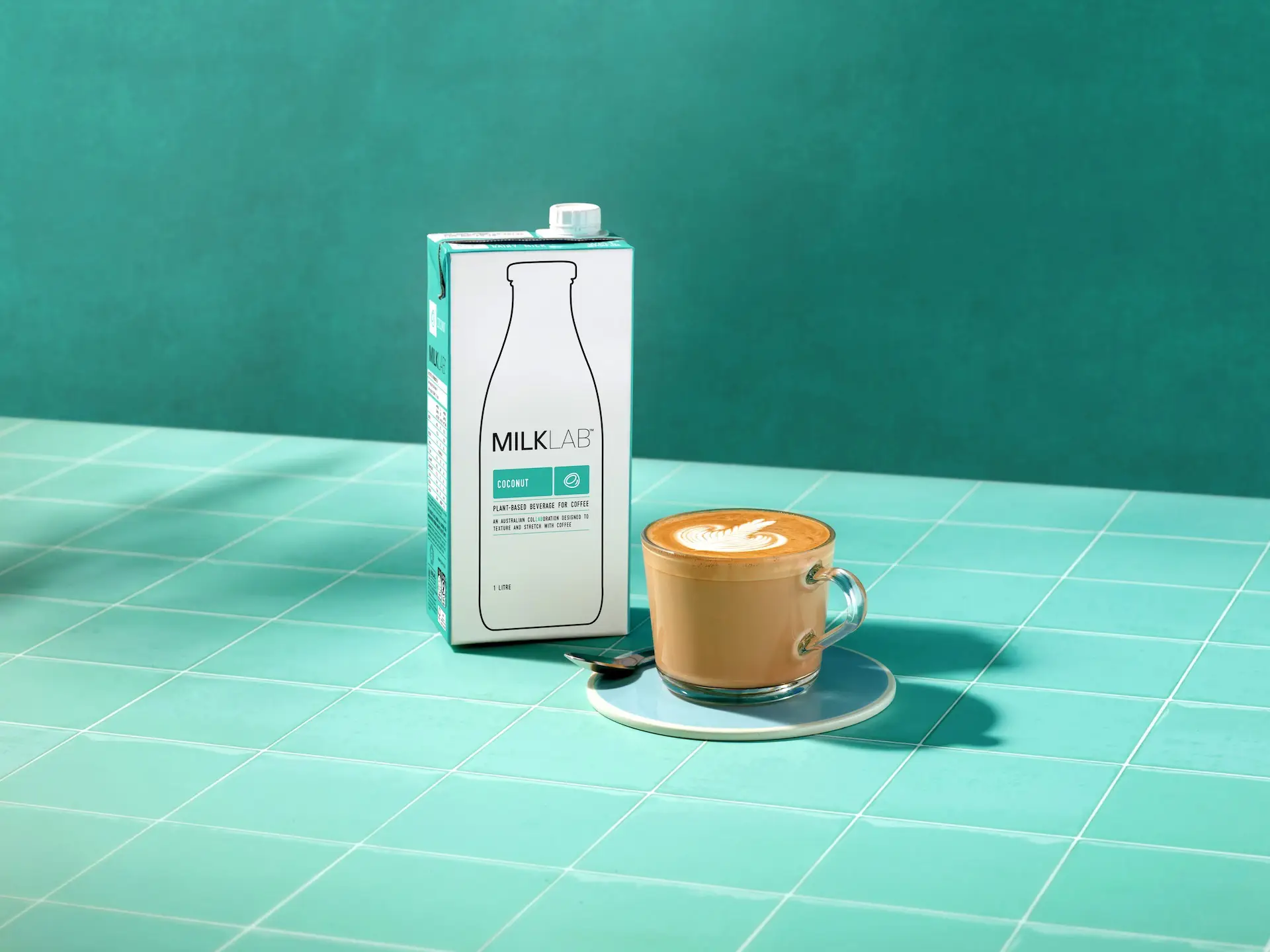 Carton of MILKLAB coconut plant-based beverage next to a glass cup of latte with leaf latte art on a turquoise tiled surface.