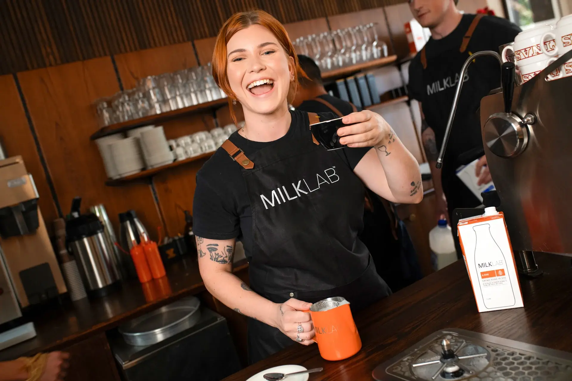 Smiling barista in a MILKLAB apron holding a black coffee cup and frothing milk in an orange MILKLAB pitcher behind a coffee counter.
