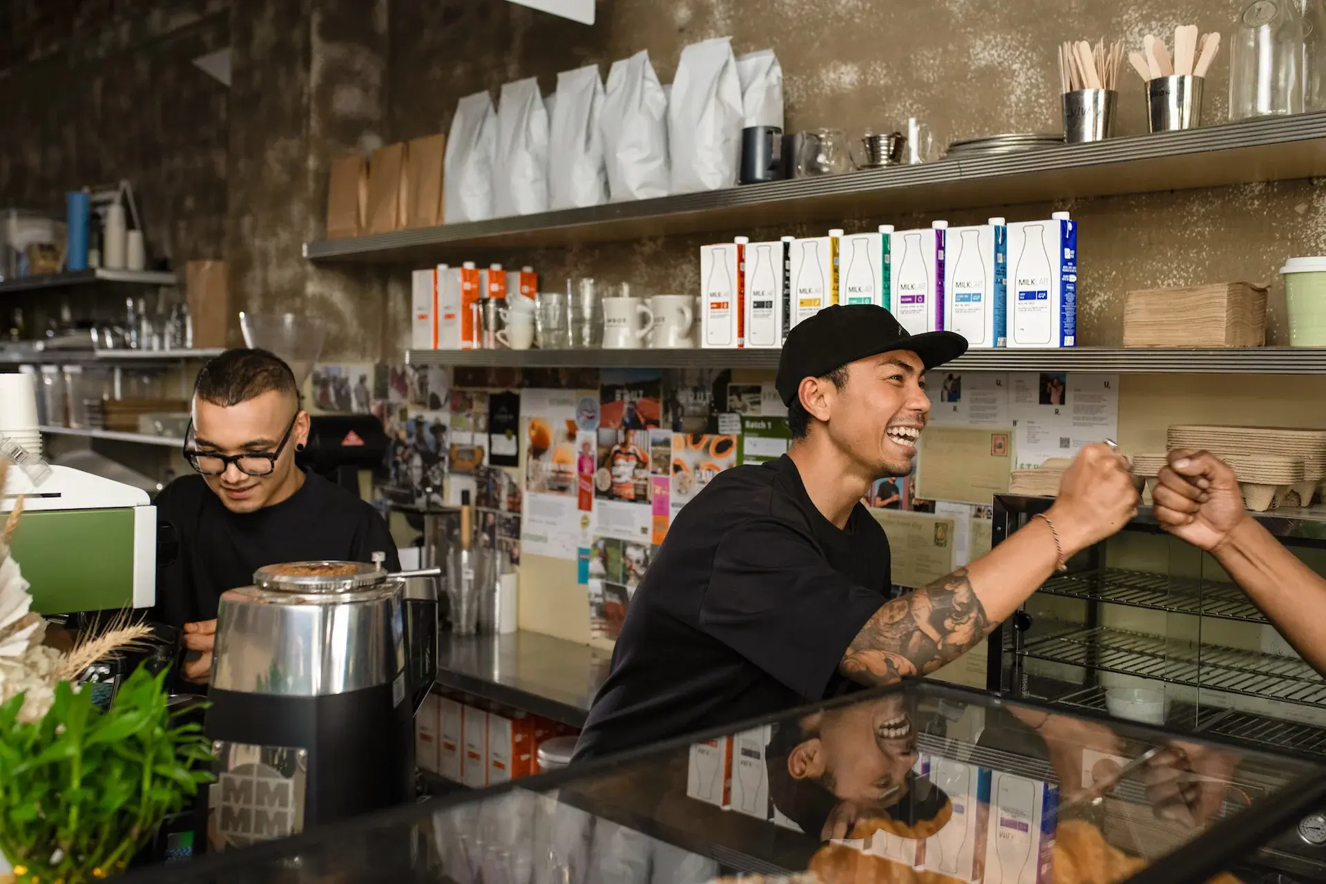 Two baristas in a coffee shop fist bumping with one smiling and another preparing a coffee machine.
