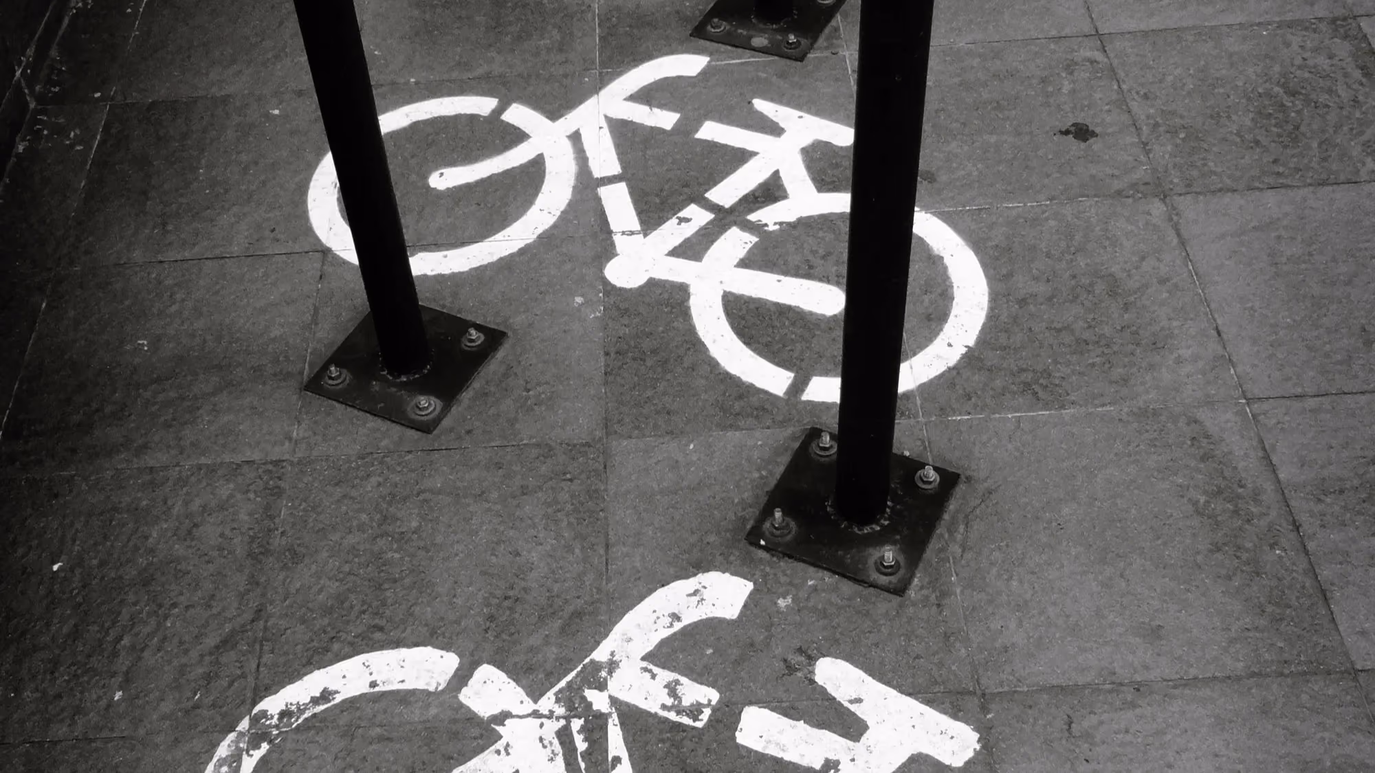 A row of metal bike racks on stone pavement, each marked with a white painted bicycle symbol, photographed in black and white.