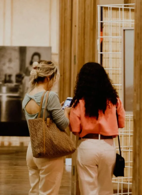 Deux femmes vues de dos dans un musée, l'une regarde son téléphone, entourées d’œuvres d'art.
