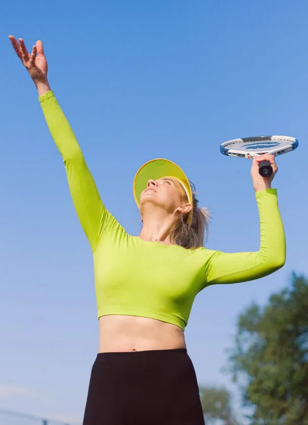 Femme en tenue de sport verte et casquette servant une balle avec une raquette de tennis sous un ciel bleu dégagé.