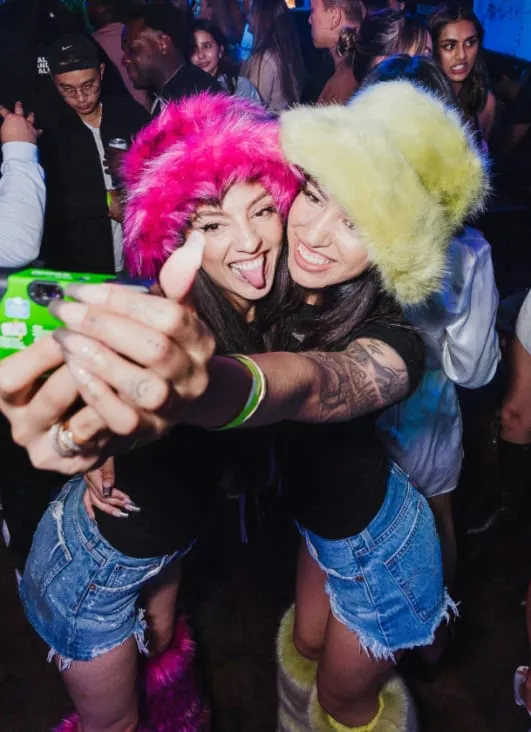 Deux jeunes femmes souriantes portant des chapeaux en fausse fourrure, des shorts en jean et des bottes assorties, prenant un selfie en soirée.