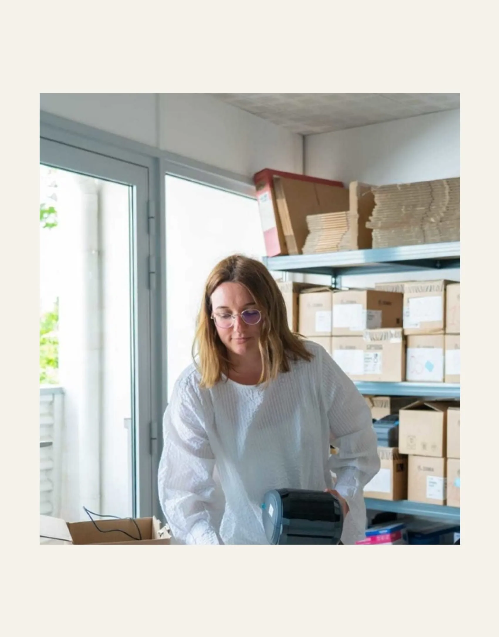 Femme avec lunettes préparant un colis dans un entrepôt avec étagères et cartons derrière.