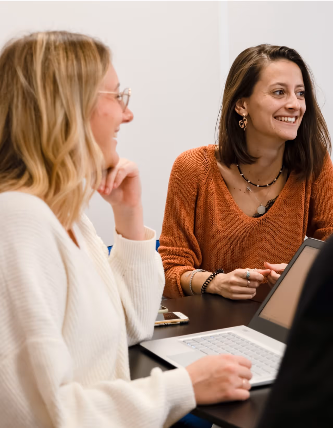 Deux femmes souriantes discutent autour d'un ordinateur portable et d'un smartphone sur une table.
