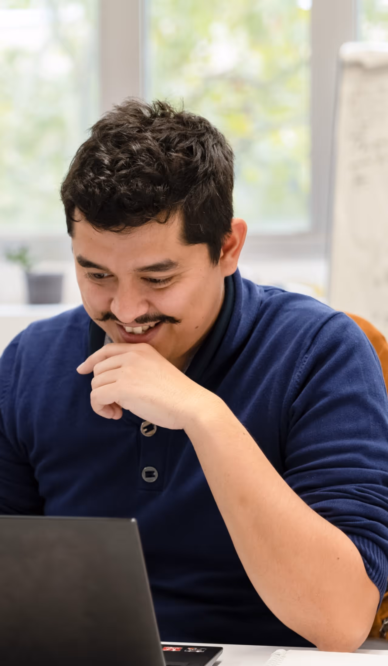 Homme souriant regardant un ordinateur portable en intérieur avec un pull bleu marine.
