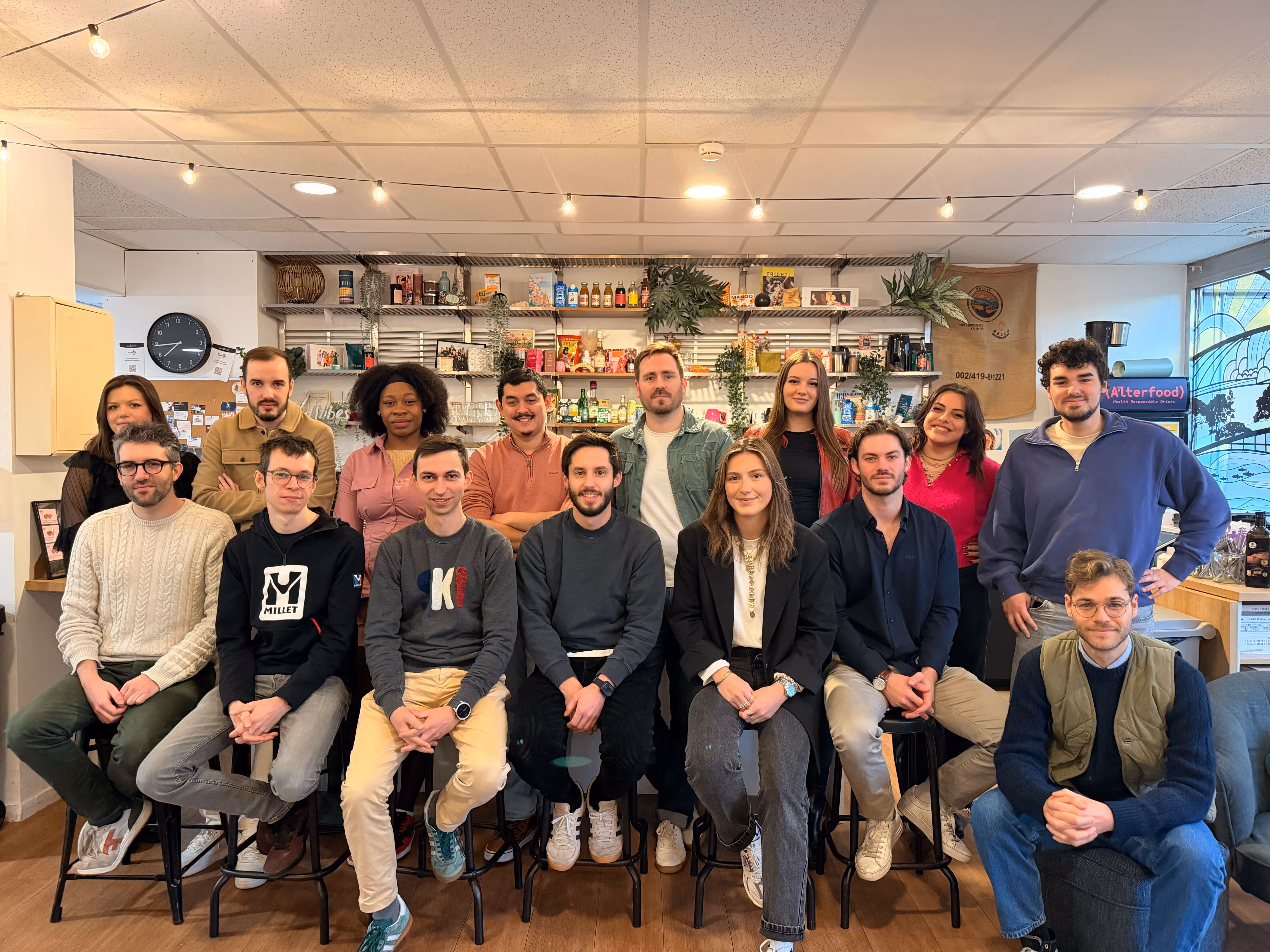 Groupe de 15 personnes souriantes posant dans un espace intérieur convivial avec étagères et lumières.