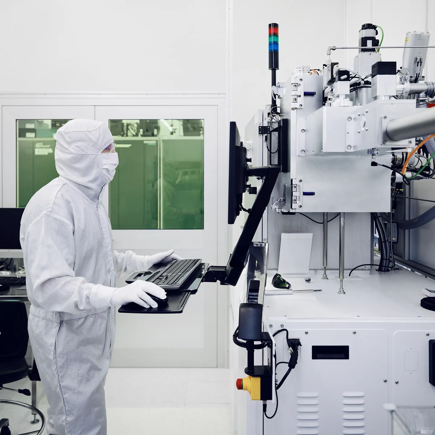 Person in a full cleanroom suit operating a computer workstation attached to industrial equipment in a sterile lab environment.
