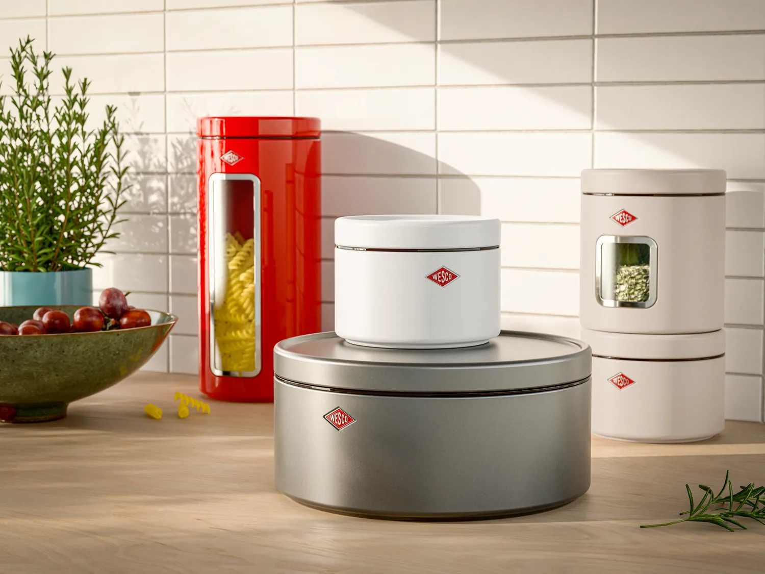 A collection of Wesco brand kitchen storage canisters displayed on a wooden countertop against a subway tile backsplash. The set includes a large brushed steel base container, white ceramic vessel, red cylindrical pasta canister, and cream-colored storage jars, each featuring the distinctive Wesco diamond logo. Styled with potted herbs, fresh produce, and natural lighting to showcase the functional yet decorative kitchenware.
