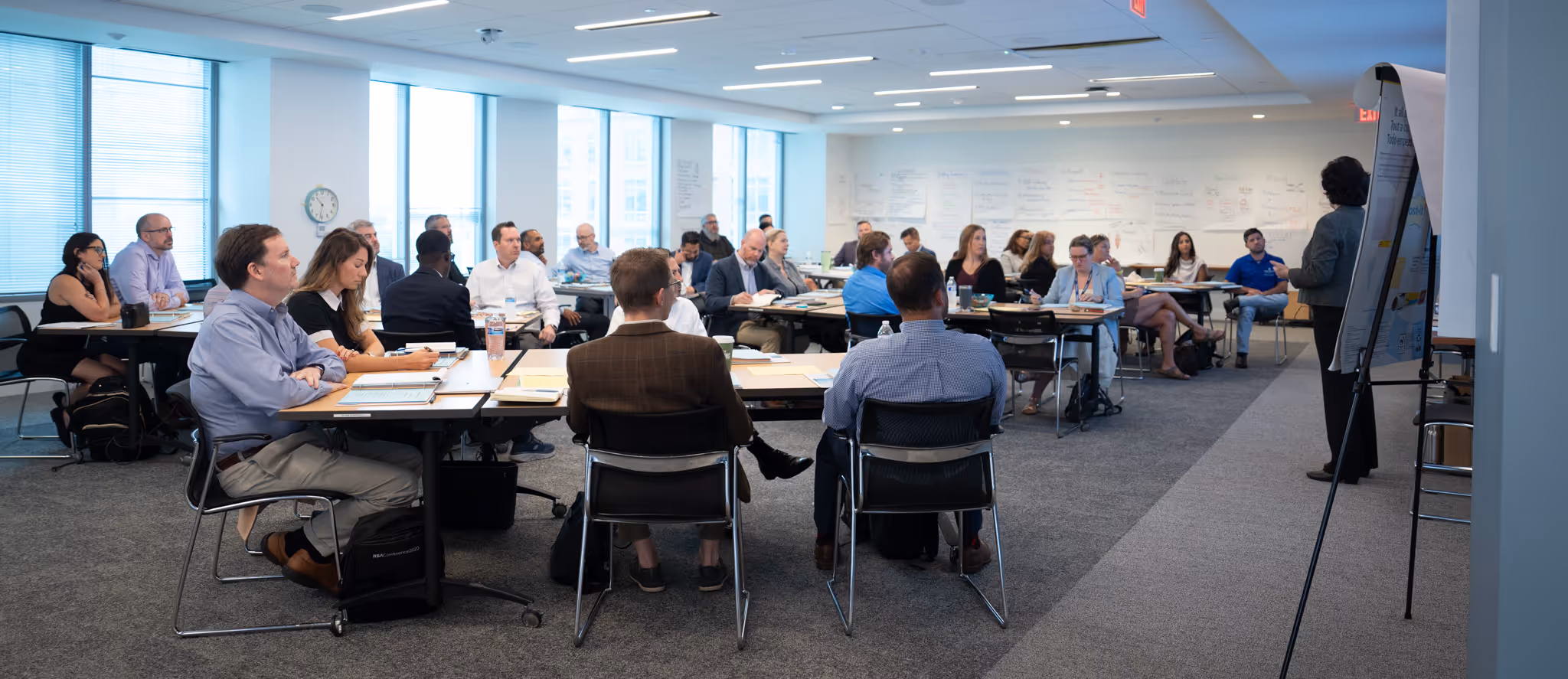 People seated around tables attentively listening to a presenter in a modern conference room with large windows and notes on the walls.