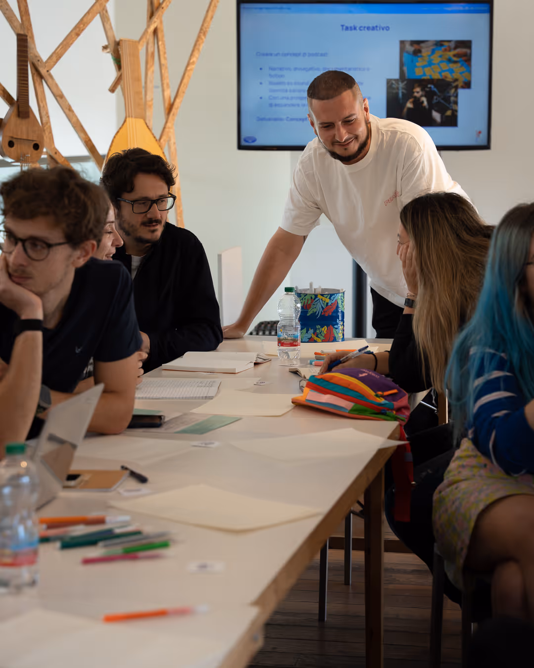 Un gruppo di persone sedute intorno a un tavolo che lavorano insieme durante un'attività creativa, con uno schermo alle loro spalle che mostra un task creativo.