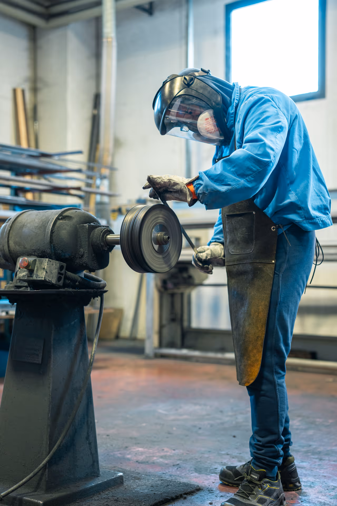 Operaio in tuta blu con maschera protettiva usa una mola abrasiva in un'officina.
