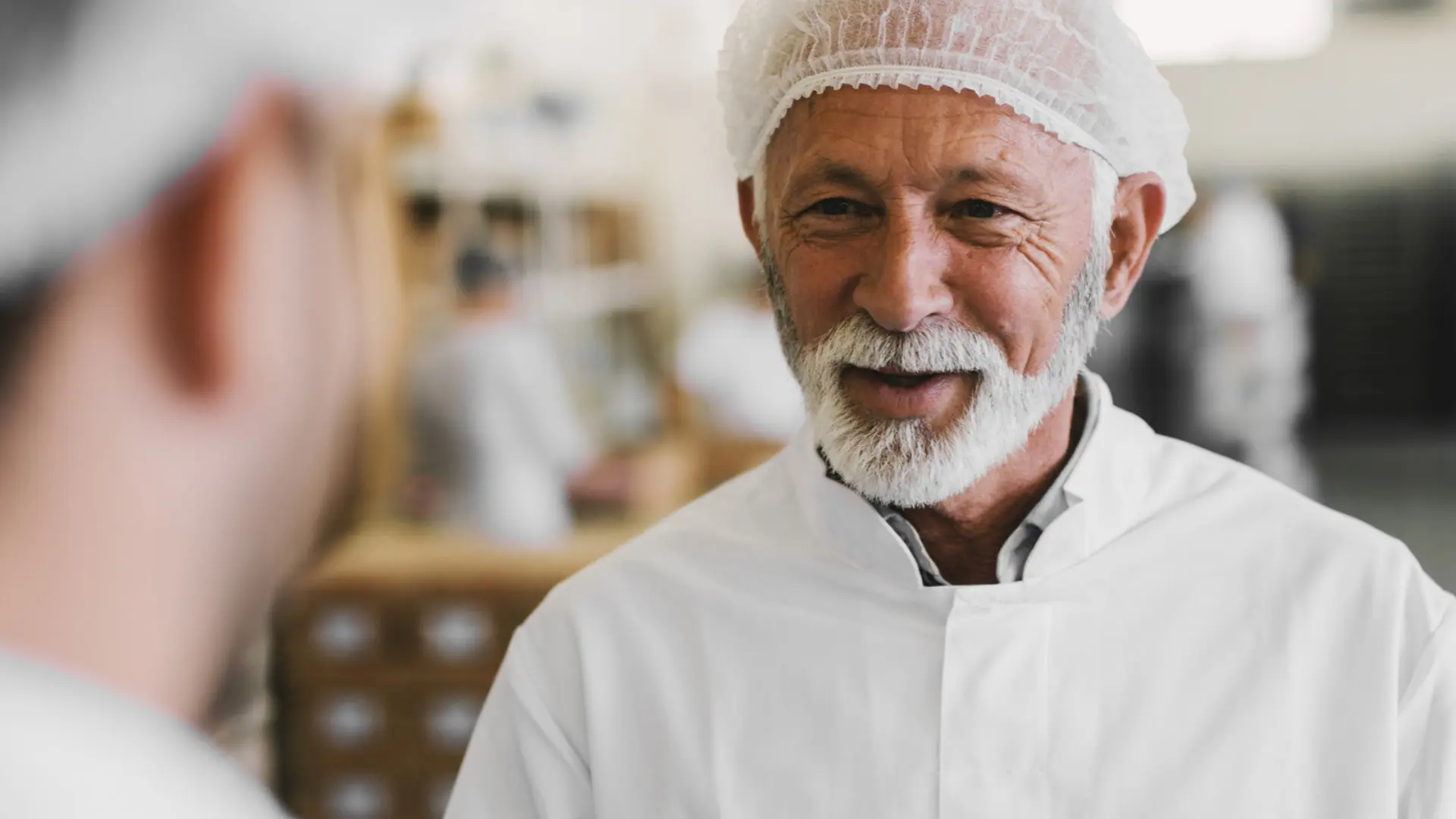 Older man with white beard and hairnet talking with a colleague in a food production facility.