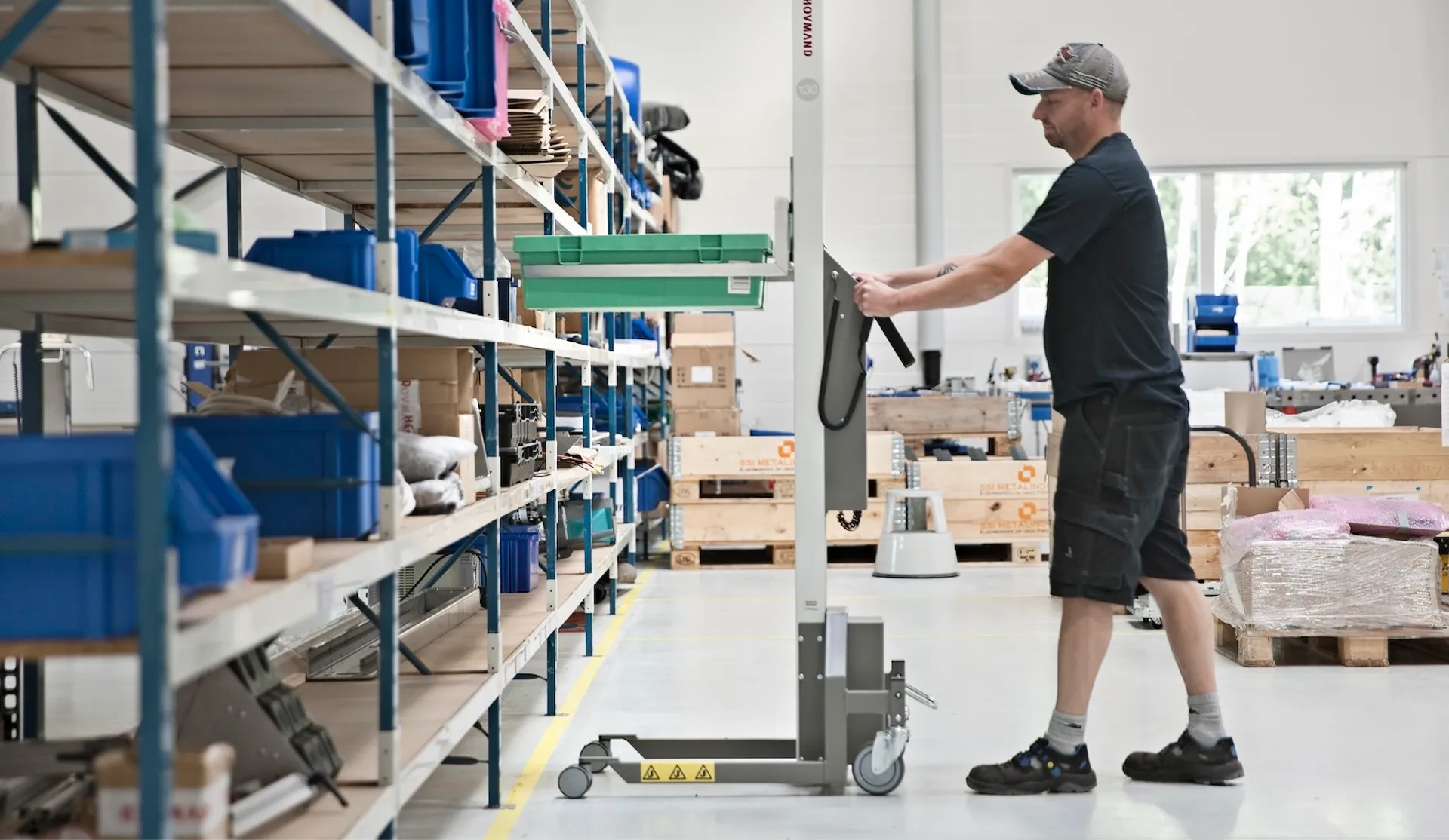 Worker in a warehouse with a hand truck next to shelves stacked with blue boxes and wooden pallets.