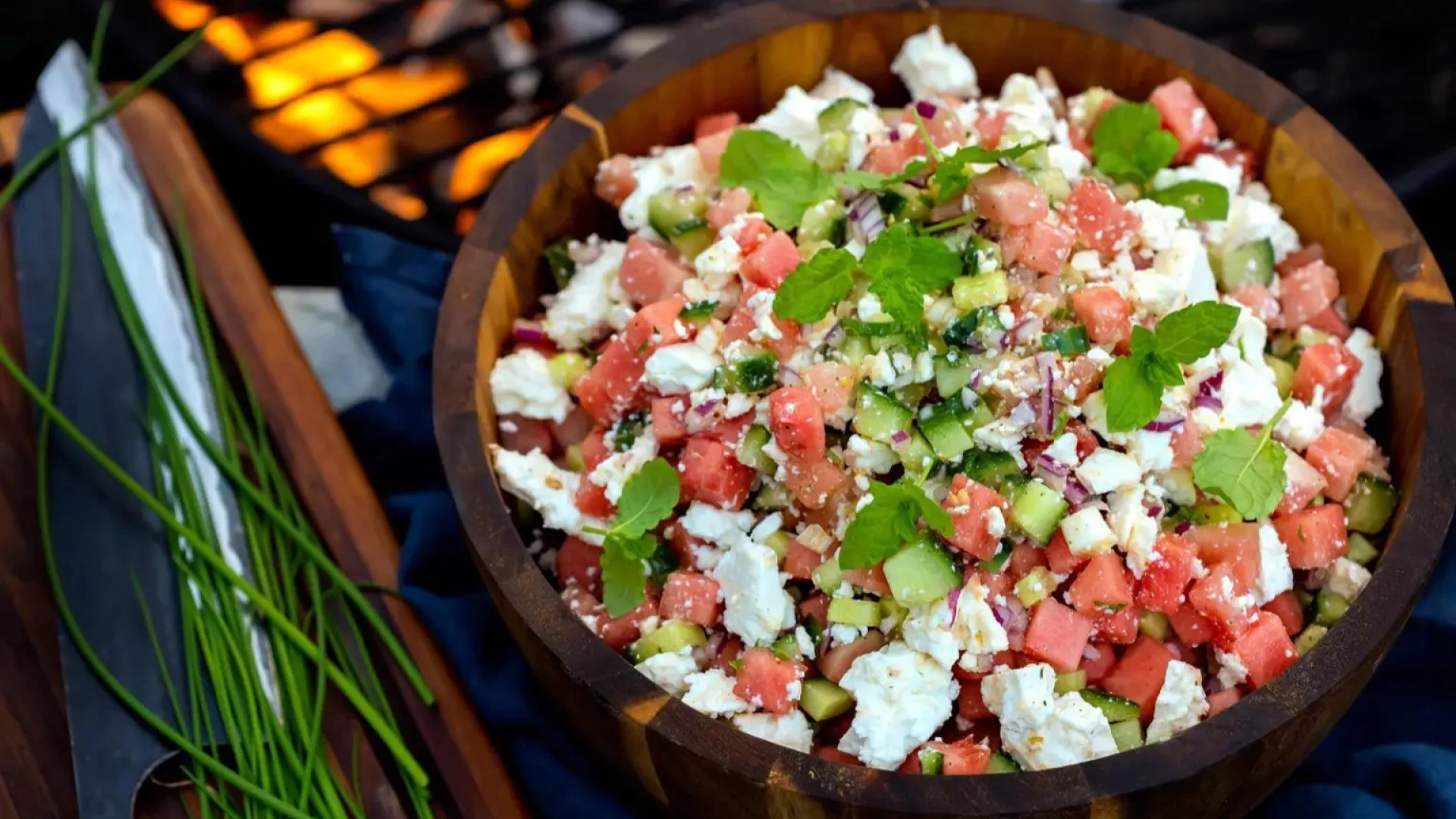 Greek salad with feta cheese, tomatoes, cucumbers and herbs in a wooden bowl near a grill.