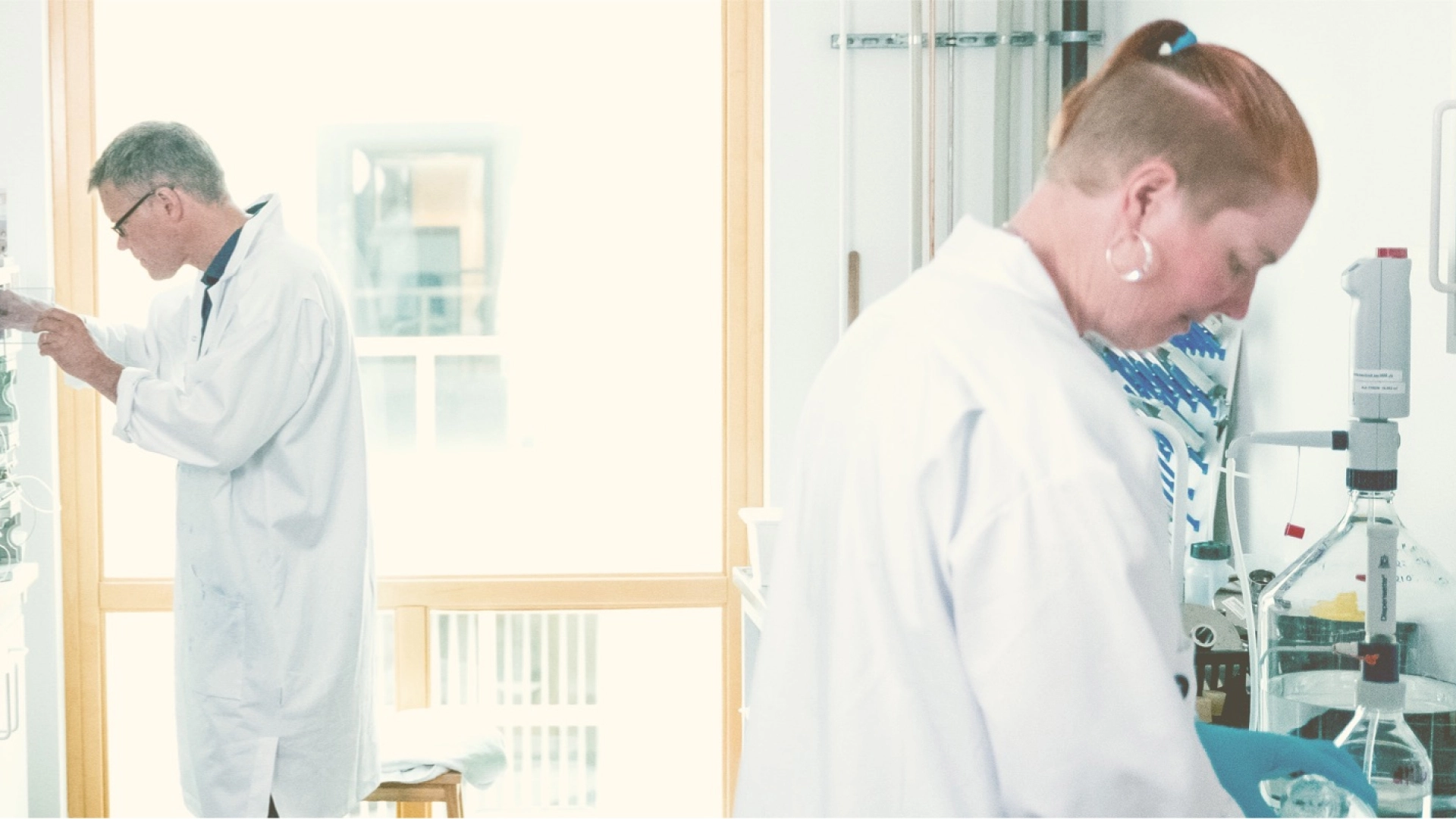 Two researchers in lab coats examining samples with laboratory equipment in a bright facility.