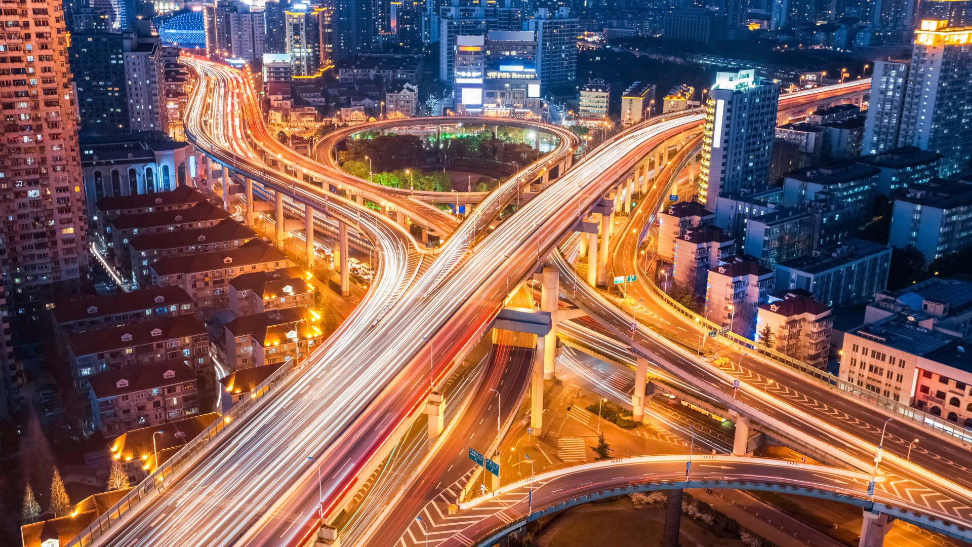 Interconnected highway interchanges with light trails at dusk, surrounded by city buildings and skyscrapers.