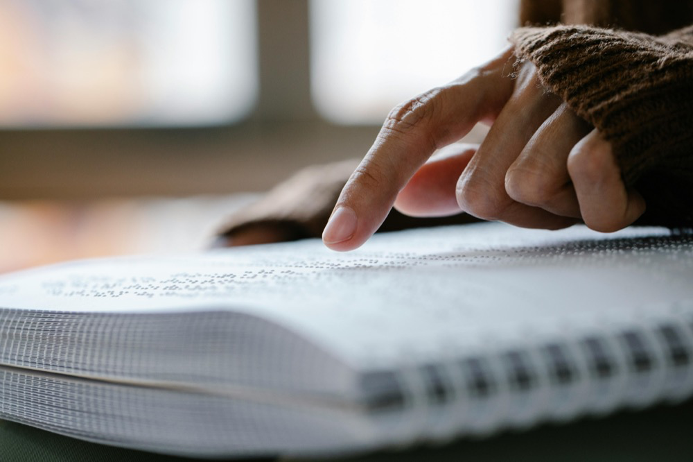 Close-up of a hand in a brown sweater reading a braille book, fingers tracing the raised dots, accessible reading material for visually impaired people.