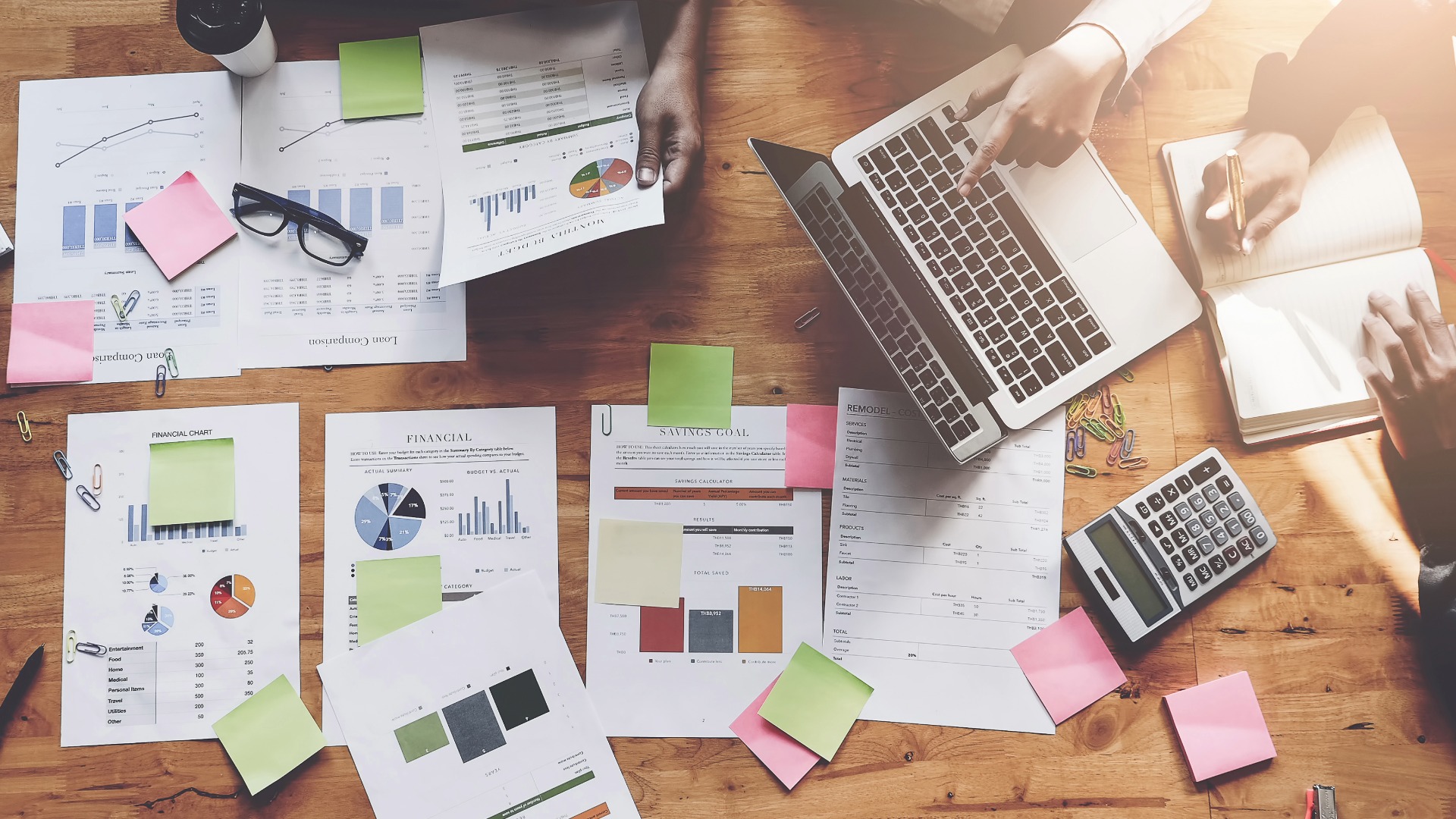 Financial documents and charts spread across a wooden table with a laptop, calculator and the hands of people working together.