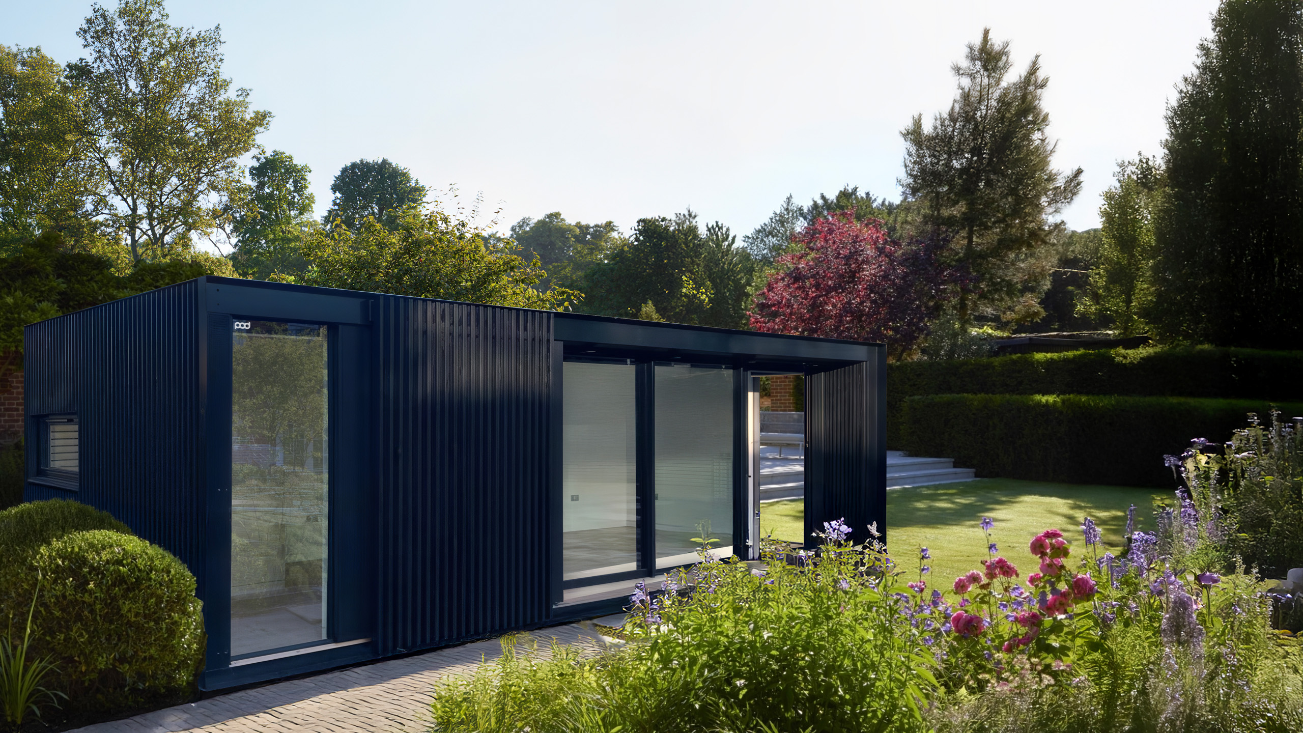 Contemporary garden building sits within flowering english summer garden, with feature black cladding and full height glazing, the building creates a modern focal point.