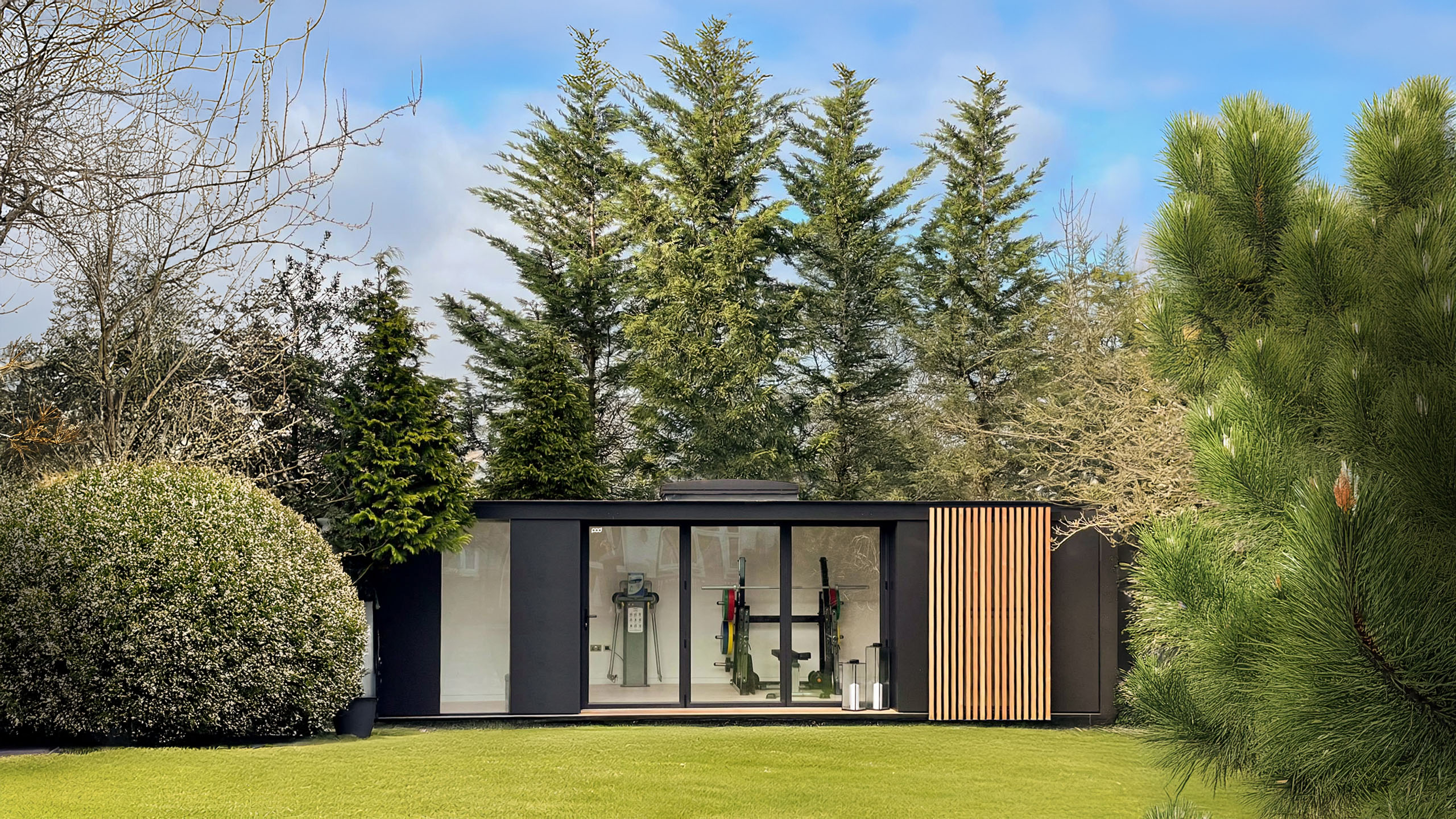 Contemporary garden building with striking black aluminium front, large glass windows and feature natural wooden cladding set amongst green lawn and tall green trees.