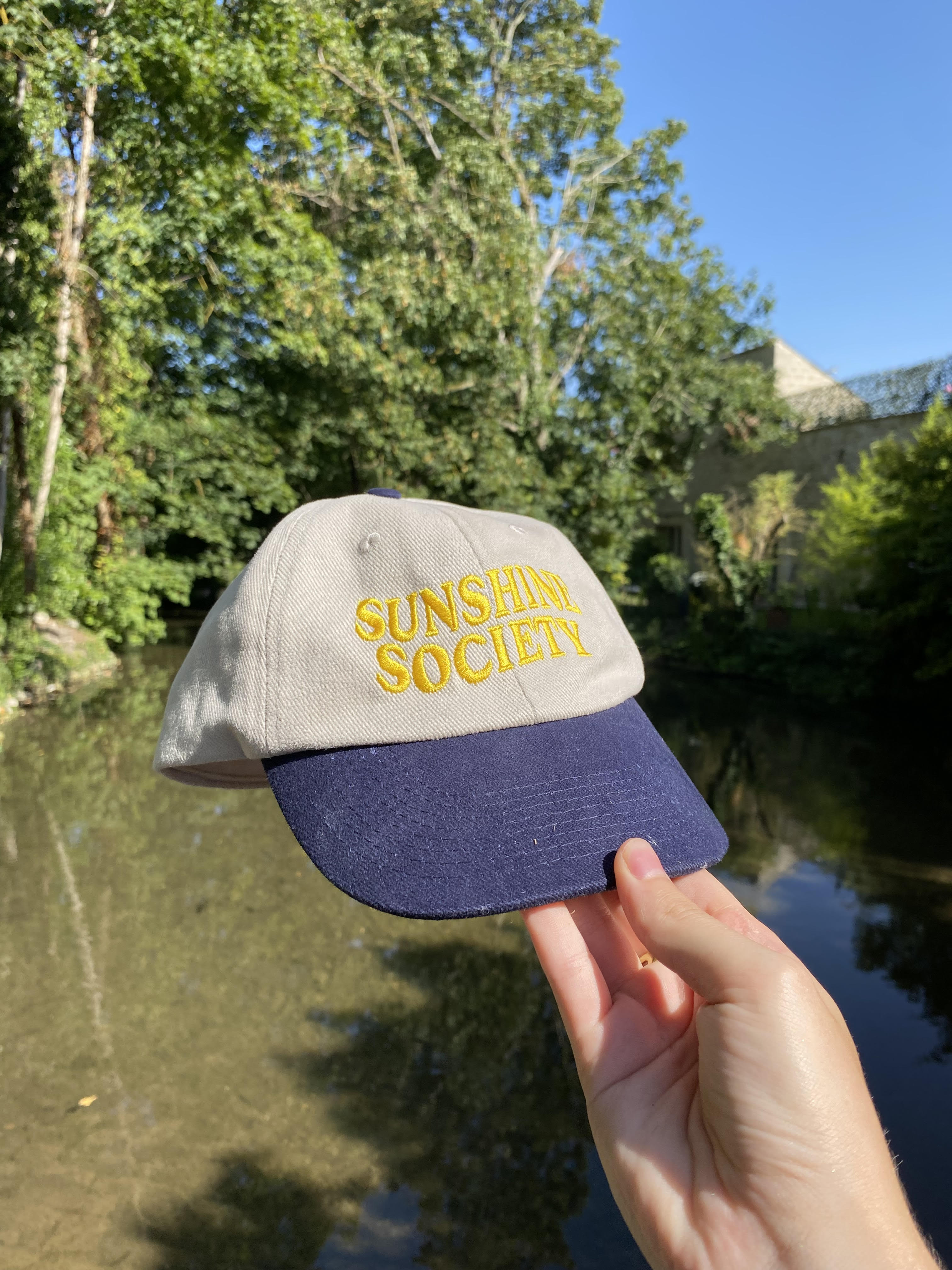 Casquette personnalisée Sunshine Society beige et marine, tenue à la main en extérieur près d’une rivière, accessoire tendance été 2025.