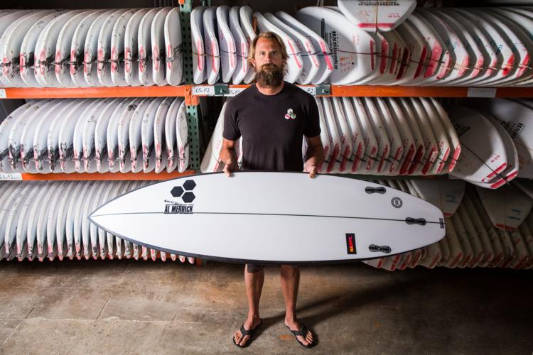 a man holding a surfboard in front of a rack of many surfboards in a surf shop