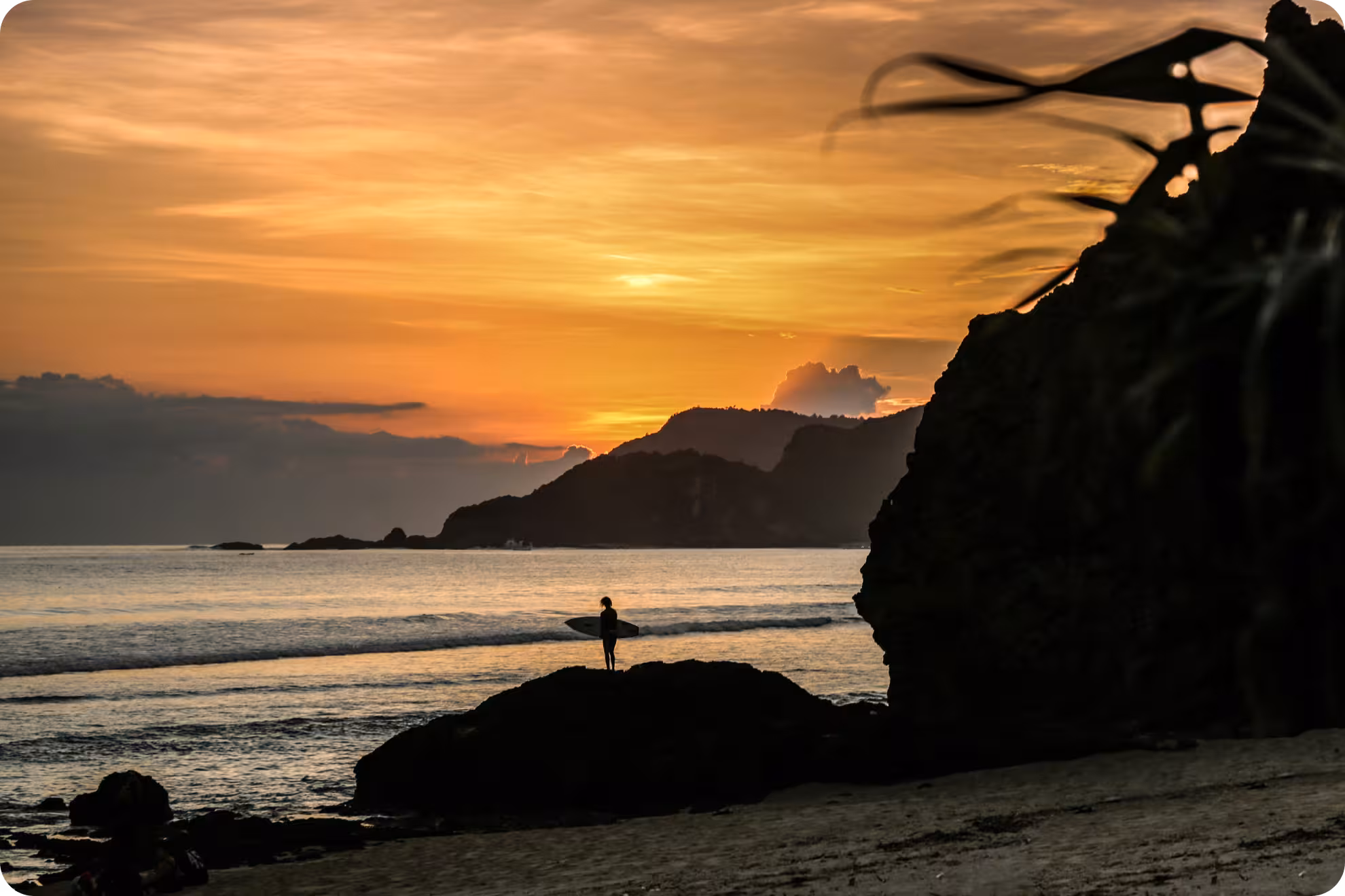 A sunset photo of Segar beach Indonesia with the silhouette of of a surfer on a rock 