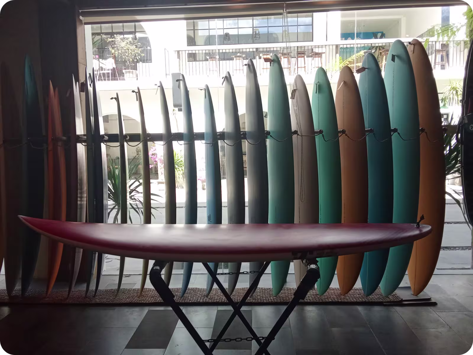 A red surfboard on a surf rack in front of a window showing more surfboards in a standing rack