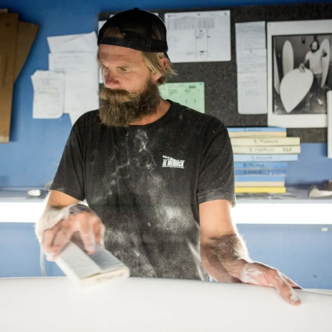 A bearded man with a backwards ball cap in a surf shop sanding a surf board