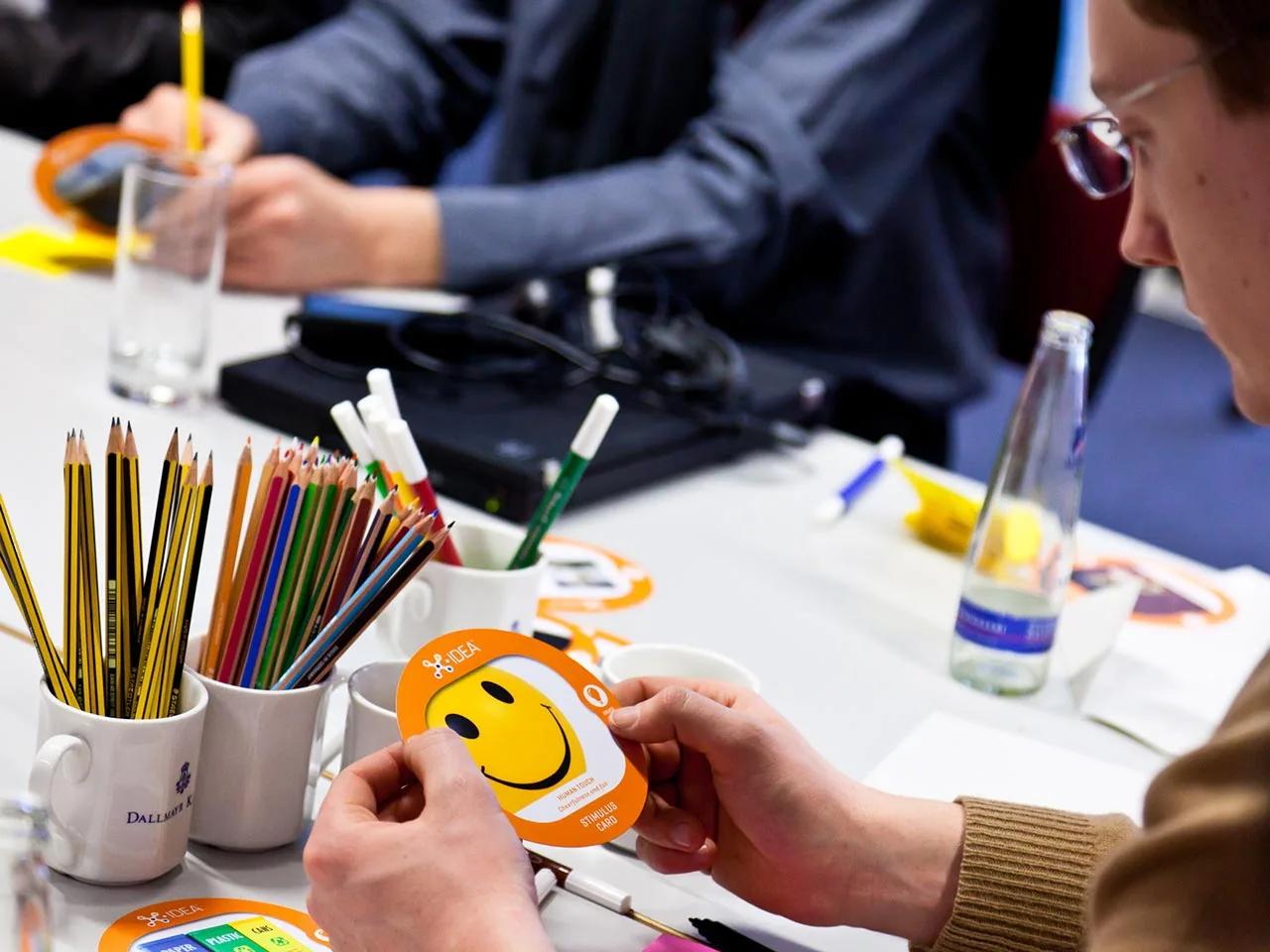 Participant holding an X-IDEA smiley stimulus card at a table with pencils, BMW project