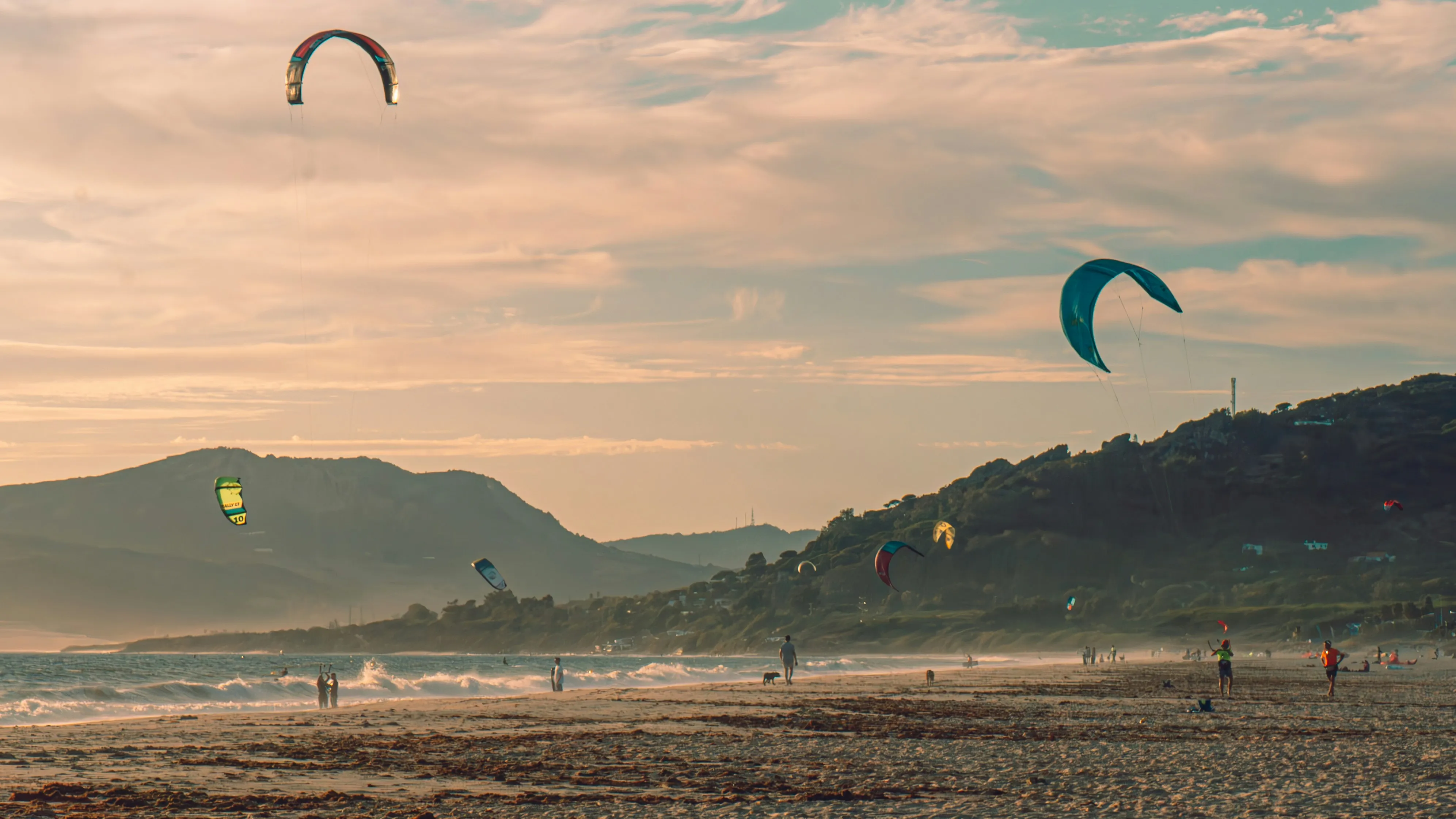 People kite surfing and walking on a sandy beach with mountains in the background at sunset.