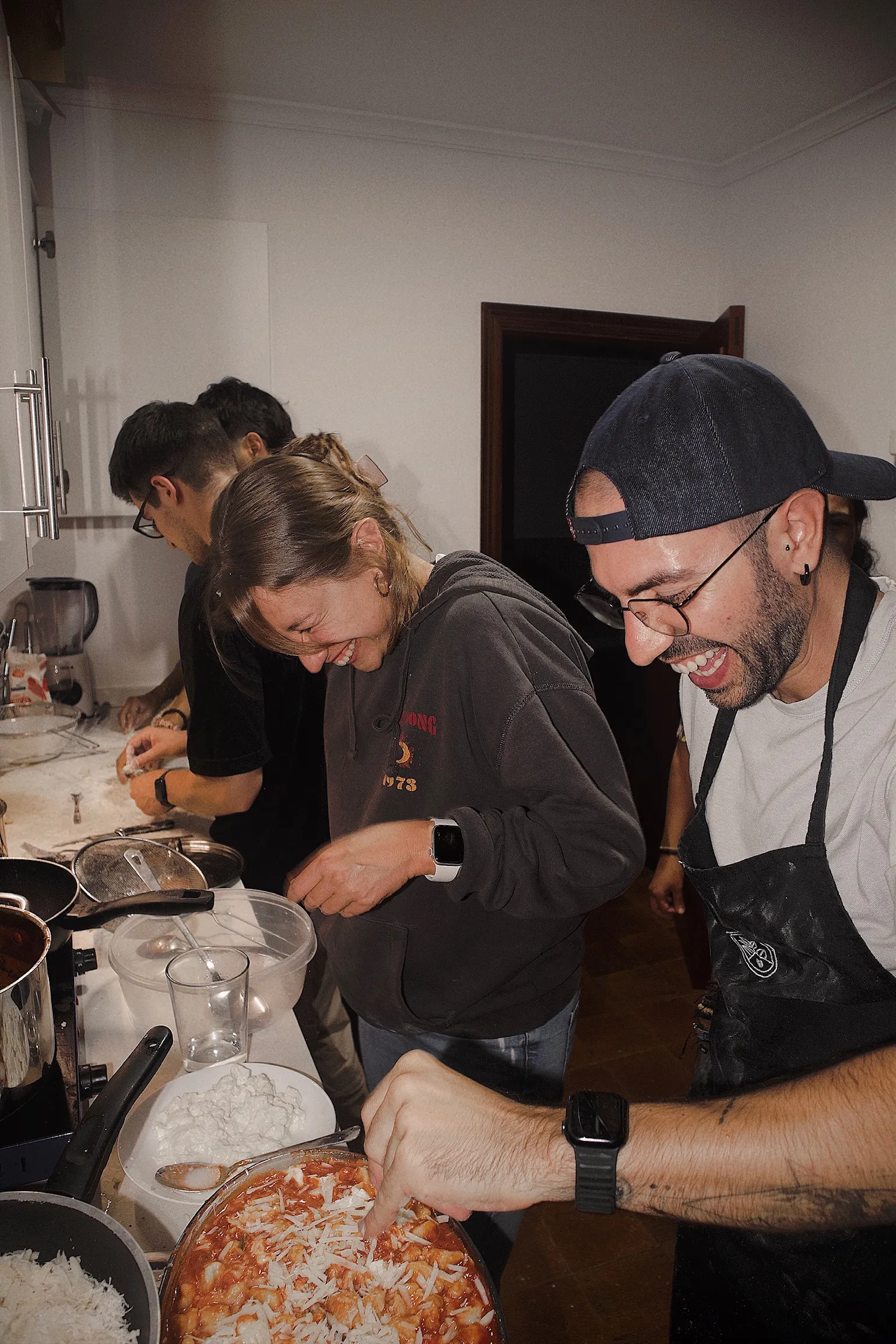A group of four people laughing and cooking together in a kitchen, one person sprinkling cheese over a dish.