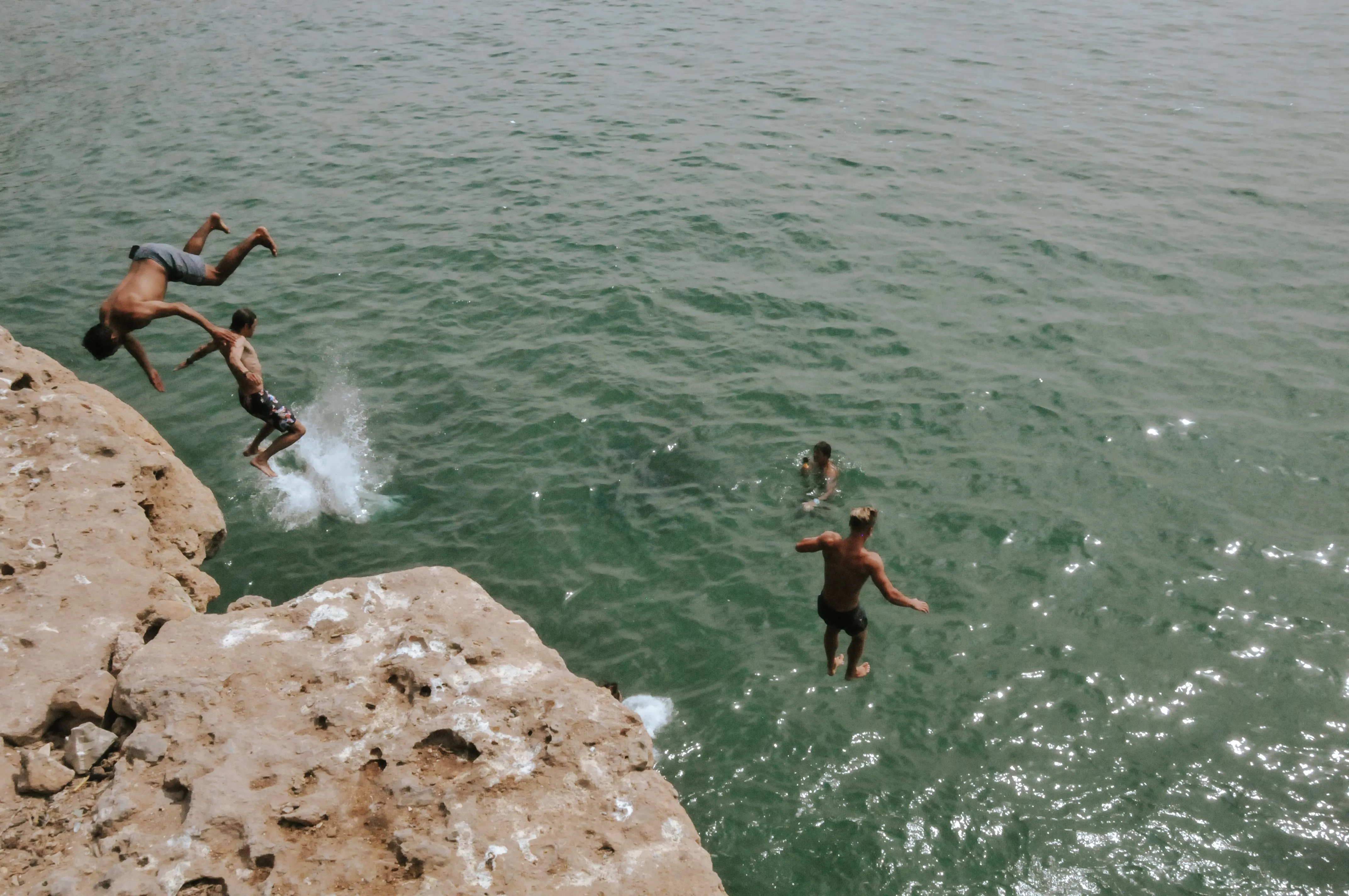 Four boys cliff jumping into green water, two in midair, one splashing into water, and one swimming.