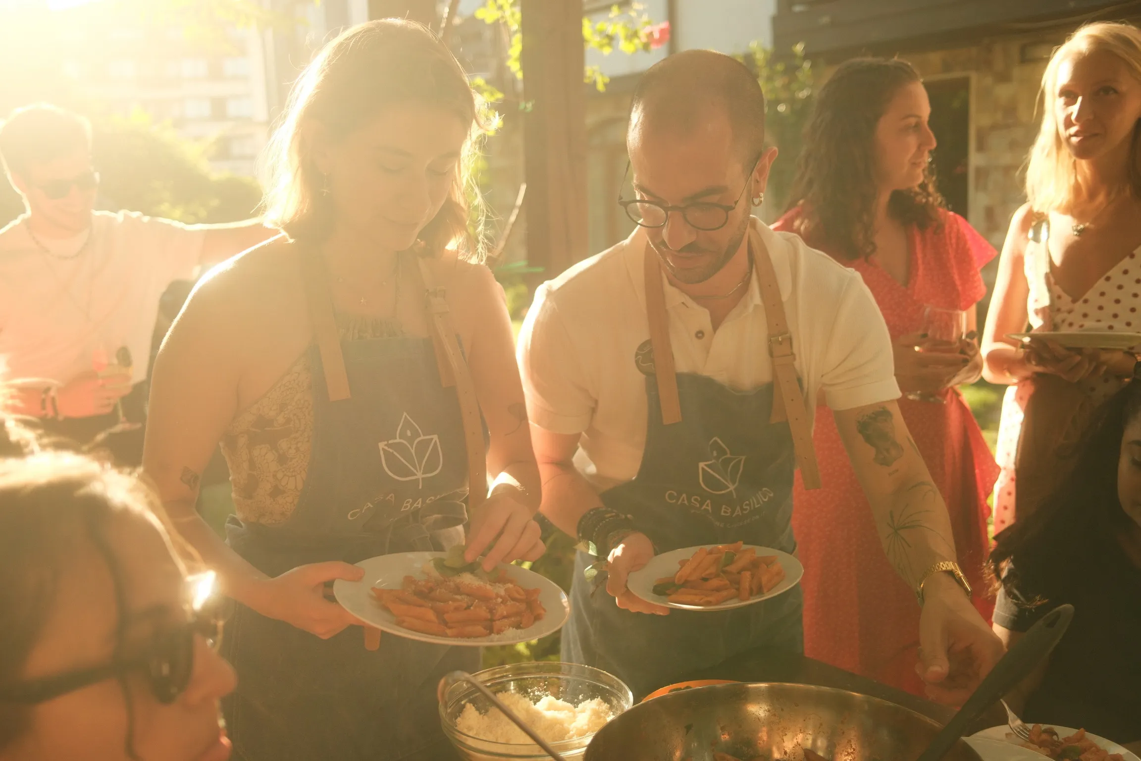 Two people wearing Casa Basilico aprons serving plates of pasta during a sunny outdoor gathering.