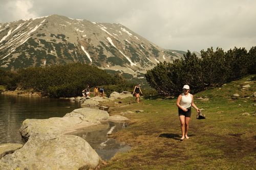 Group of hikers walking barefoot by a calm lake near mountain shrubbery with a large snow-dotted mountain in the background.
