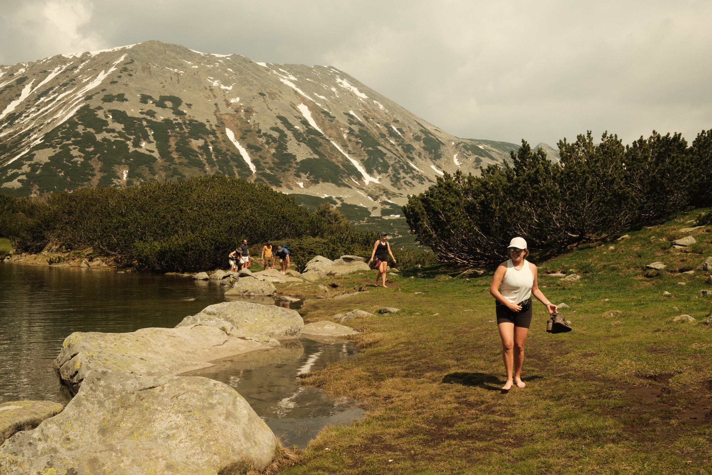 Group of hikers walking barefoot by a calm lake near mountain shrubbery with a large snow-dotted mountain in the background.