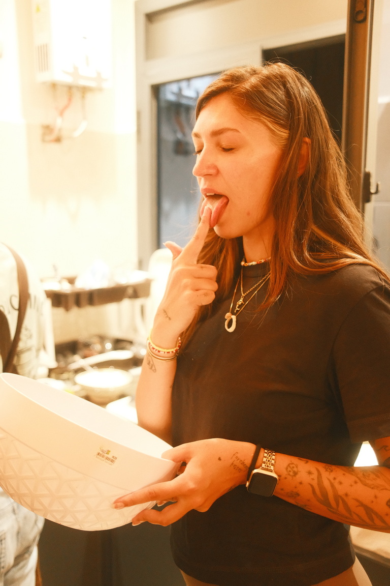 Young woman tasting food with her finger from a white bowl she is holding in a kitchen.